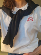 Person wearing a white t-shirt with a red embroidery of a pizza and a black jumper over their shoulder.