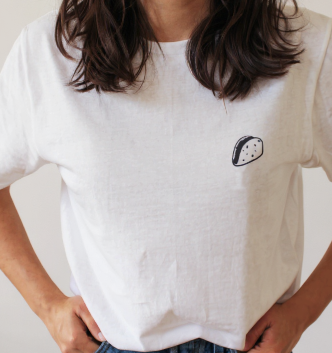 Person wearing a white t-shirt with a small black design of a taco embroidery on a plain background