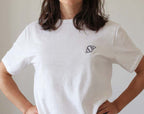 Person wearing a white t-shirt with a small black embroidery of an croissant on a plain background