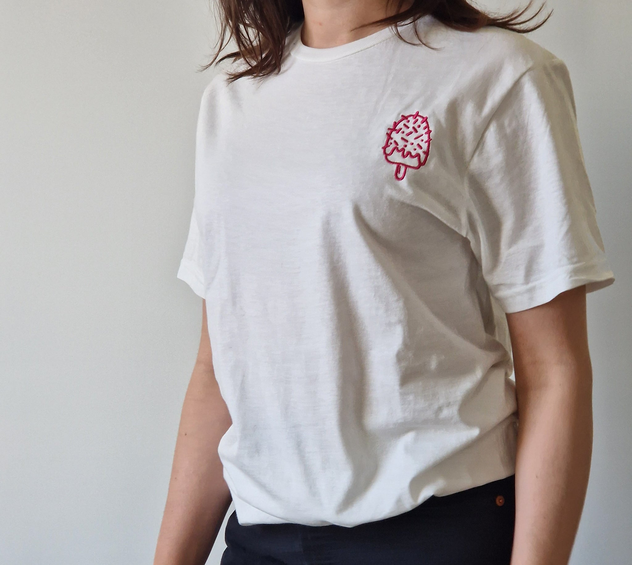 Person wearing a white t-shirt with a pink ice cream cone embroidery on a plain background